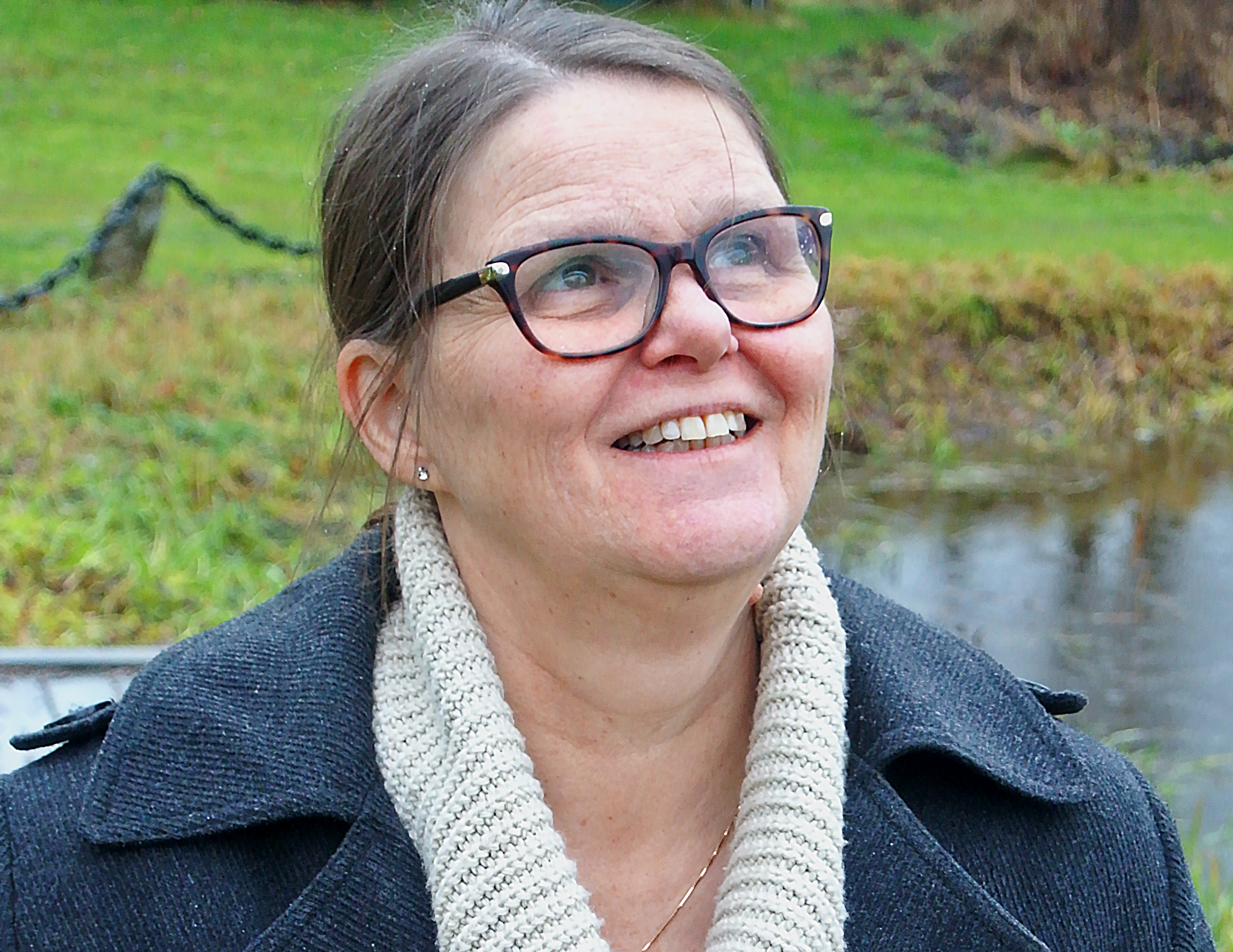 En kvinna ler med tänderna och tittar upp mot himlen. Hon har en grå kappa, beige krage, glasögon och uppsatt brunt hår. Bakom skymtar grönt gräs och ett vattendrag.