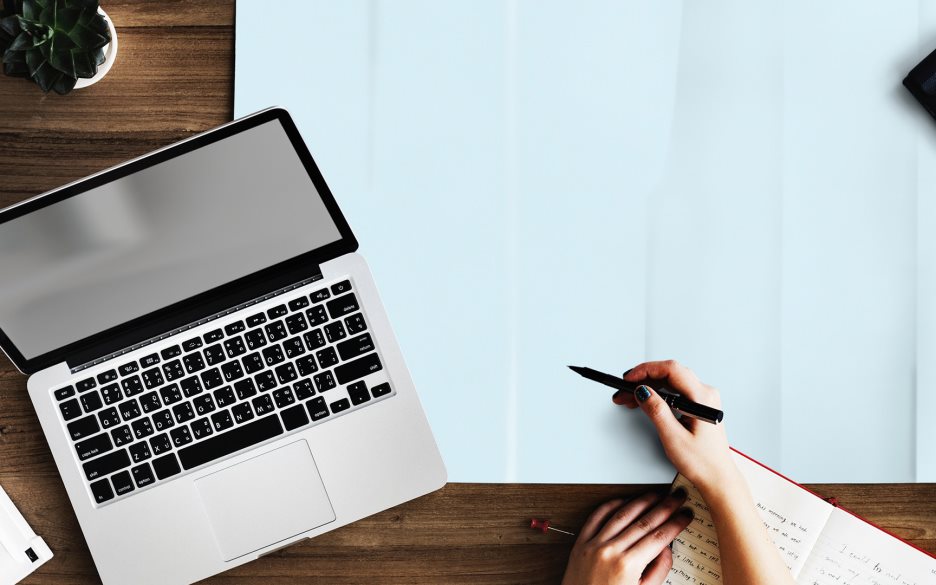 Bild ovanifrån på en dator, en kaffekopp, en hand som håller i en penna och en skrivbok.