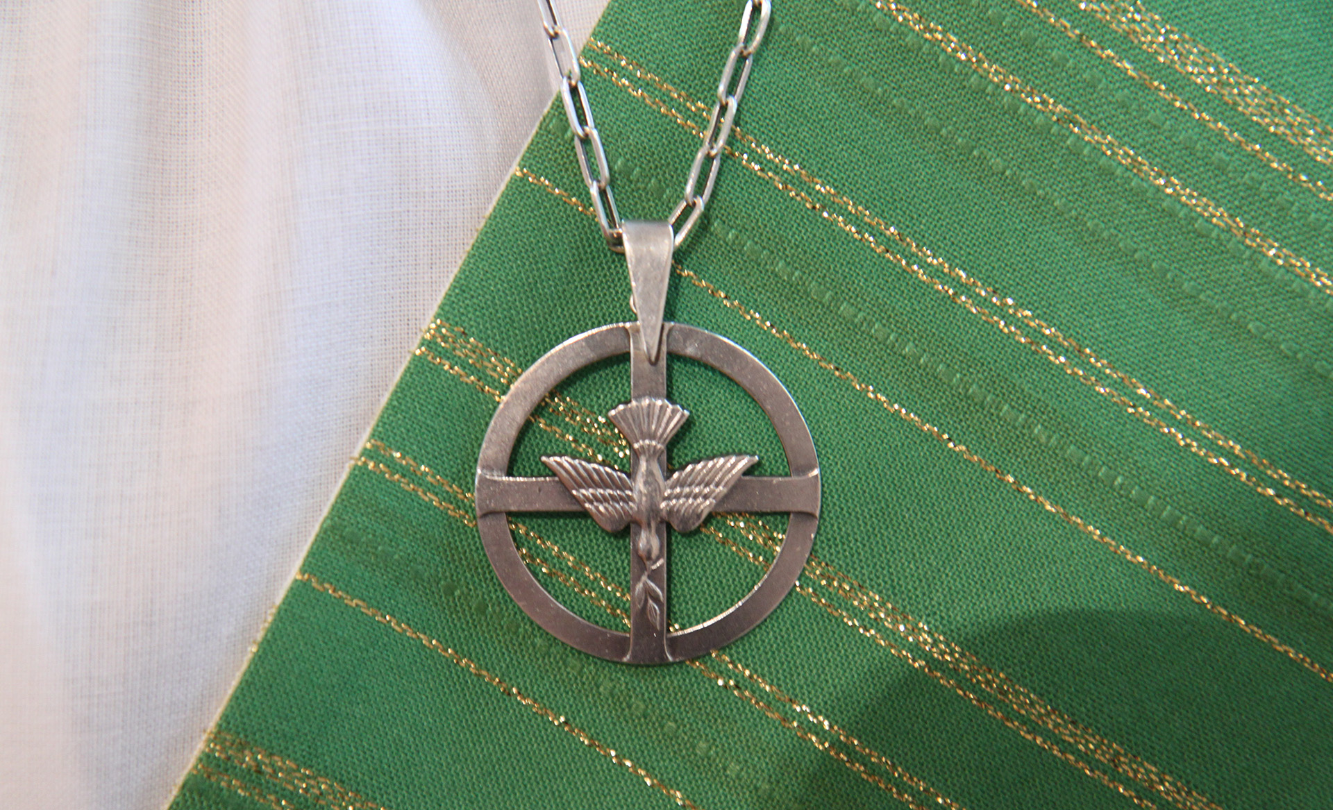 Diakonsmycke i silver som hänger mot en stola i grönt med guldtrådar.