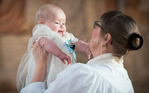 Präst som håller upp en leende bebis i dopklänning.