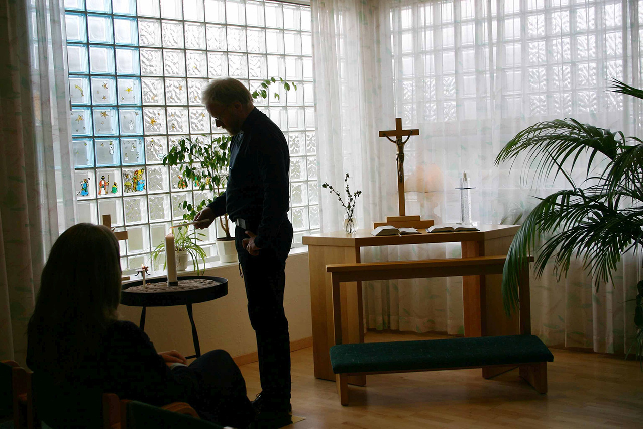 En man står vid en ljusbärare i ett andaktsrum, bredvid honom är ett altare med ett kors.