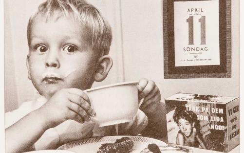 Äldre bild på liten pojke som äter frukost och det står en fastebössa på bordet. 