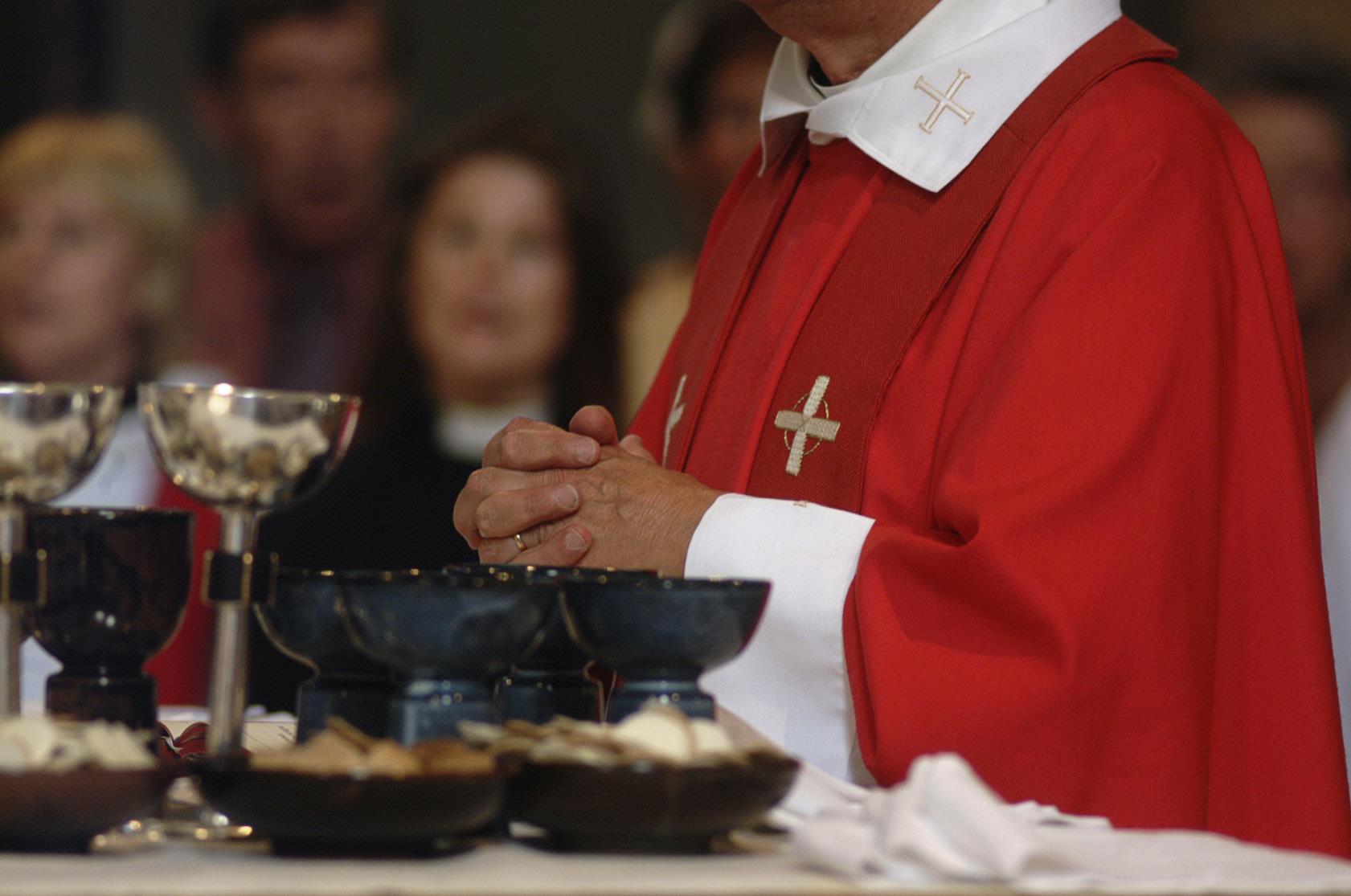 En biskop i röd mantel håller händerna knäppta framför ett bord fyllt av kalkar och skålar med oblat.