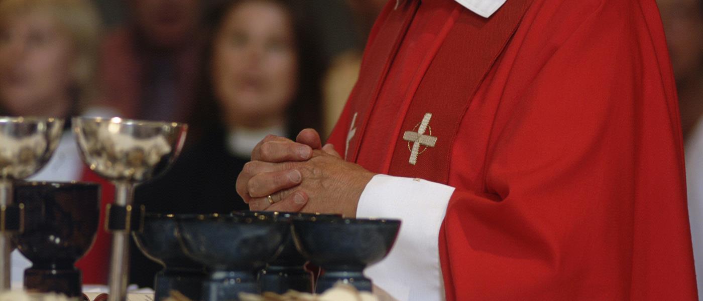 En biskop i röd mantel håller händerna knäppta framför ett bord fyllt av kalkar och skålar med oblat.