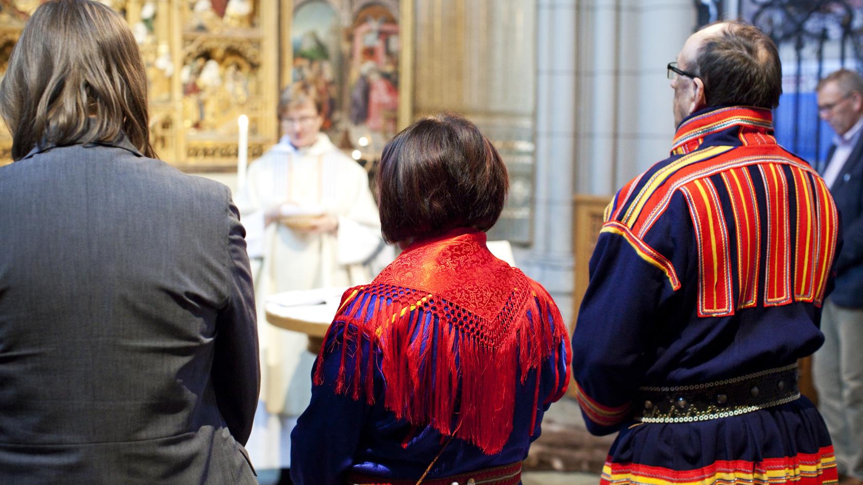 Tre personer varav två klädda i samiska folkdräkter står längst fram vid en gudstjänst i kyrkan.