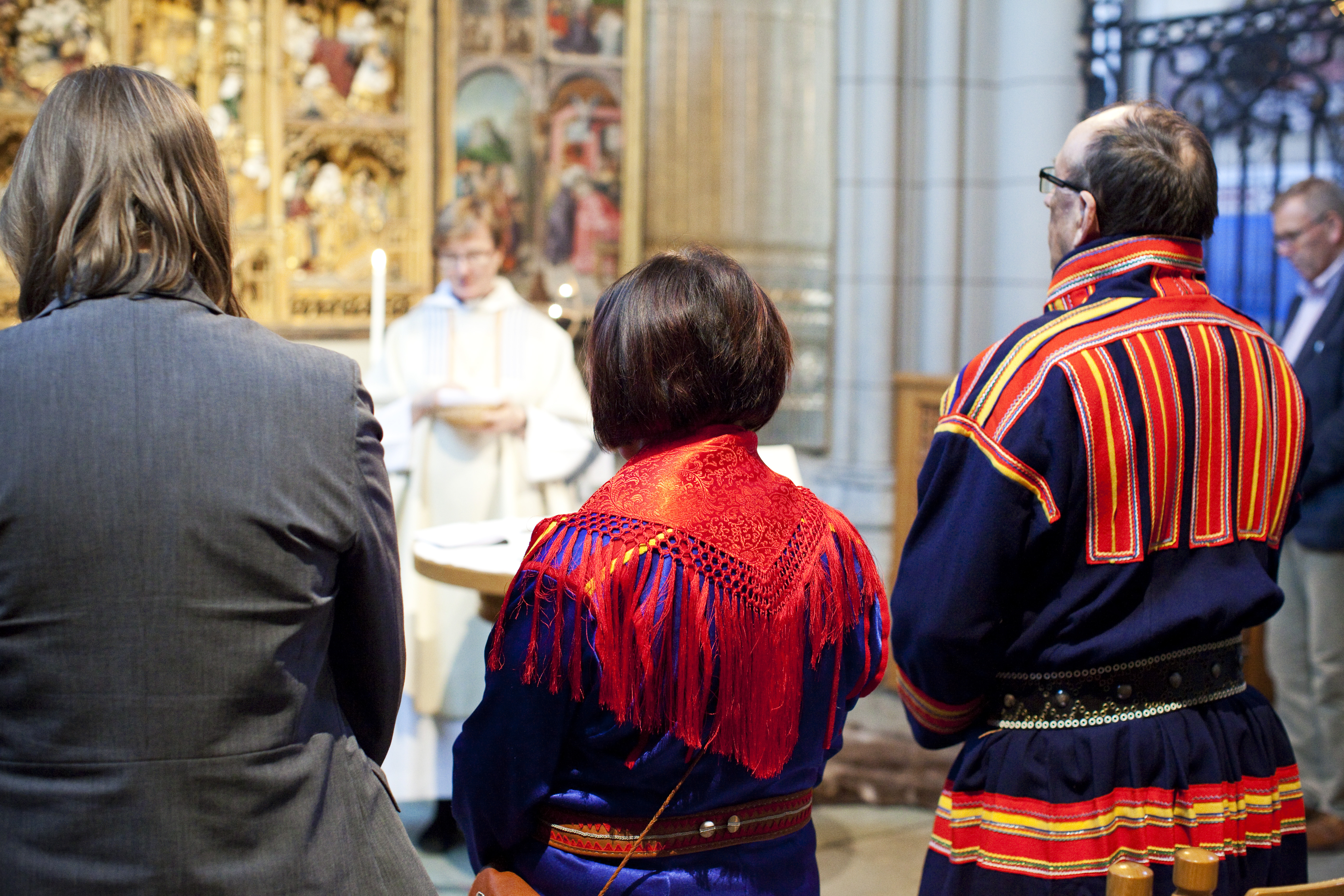 Tre personer varav två klädda i samiska folkdräkter står längst fram vid en gudstjänst i kyrkan.