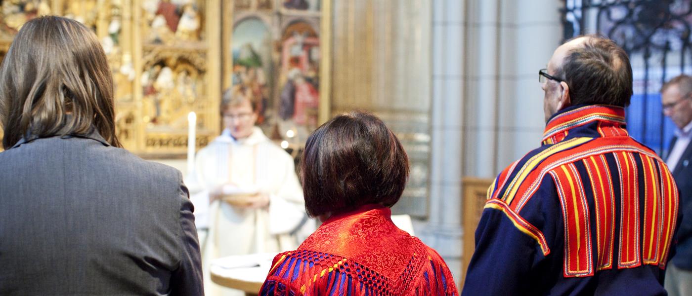 Tre personer varav två klädda i samiska folkdräkter står längst fram vid en gudstjänst i kyrkan.