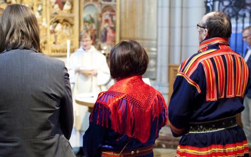 Tre personer varav två klädda i samiska folkdräkter står längst fram vid en gudstjänst i kyrkan.