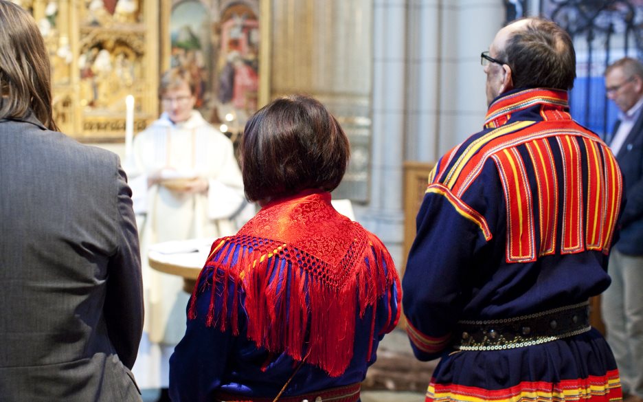 Tre personer varav två klädda i samiska folkdräkter står längst fram vid en gudstjänst i kyrkan.