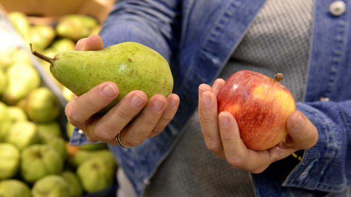 Någon står i mataffären och håller ett päron i ena handen och ett äpple i andra.
