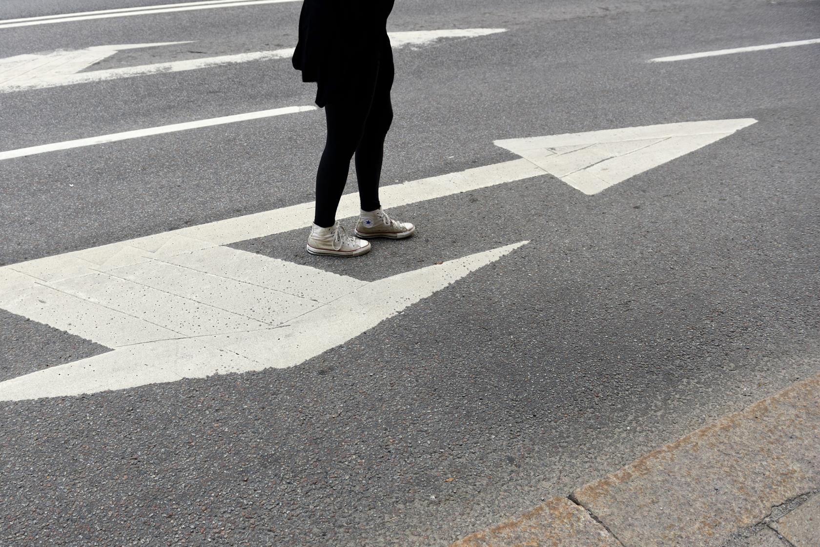 En ung person står på en körriktningsvisare målad på asfalten. En pil pekar framåt och en annan åt sidan.
