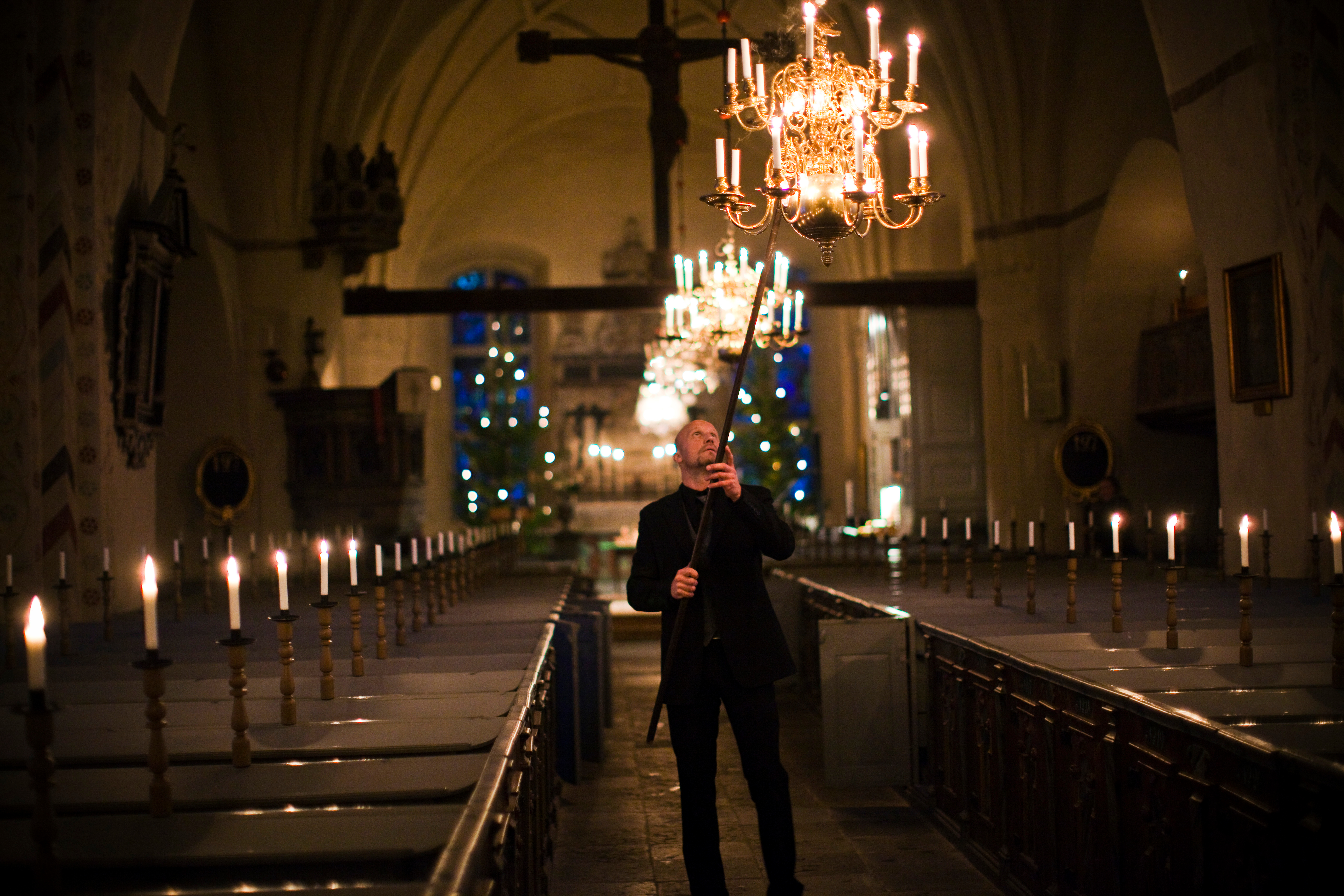 En man i en kyrka använder en lång pinne för att tända ljuskronorna i taket.