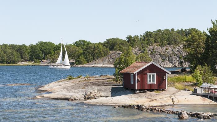 En liten röd stuga på en klippa vid vattnet. En segelbåt i bakgrunden.