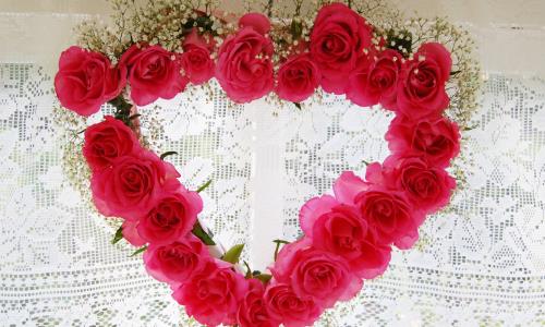 En blomsterkrans i form av ett hjärta av rosa rosor hänger vid en spetsgardin.
