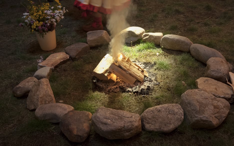 Någon med en samisk klänning och samiska skor står vid en eldstad med liten brasa och en hink med sommarblommor.