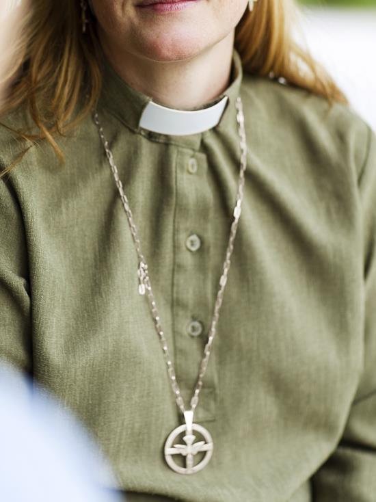 En kvinnlig diakon klädd i grön frimärksskjorta och med diakonemblemet hängande runt halsen i en lång kedja.