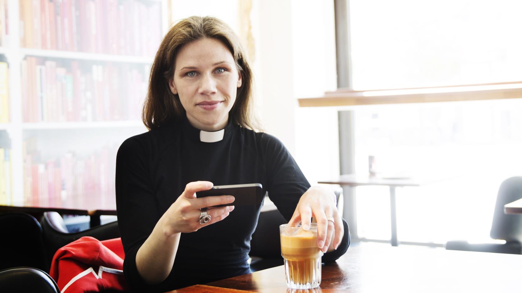 En kvinnlig präst sitter inomhus vid ett bord med en kaffe och mobiltelefonen i handen.