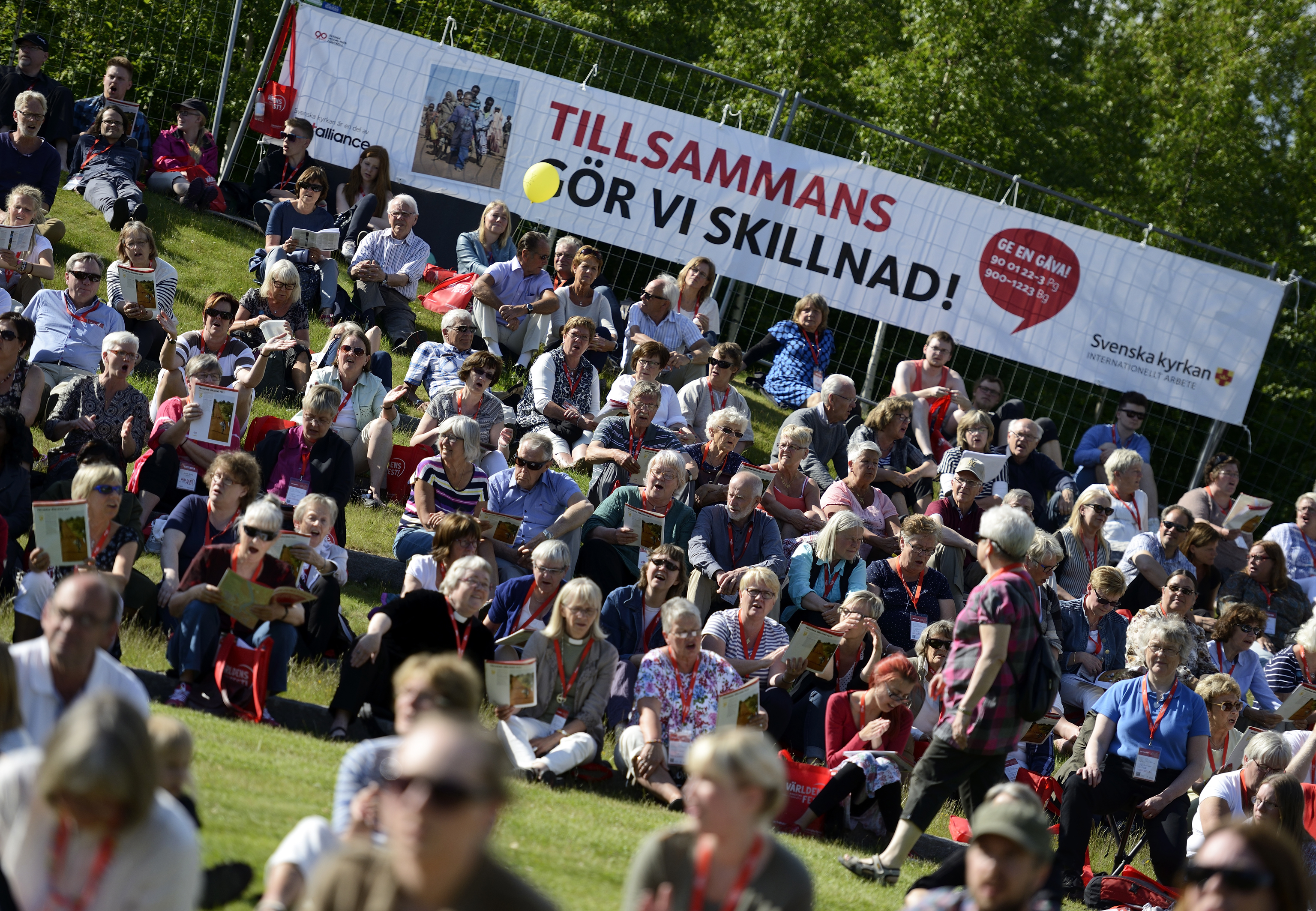 En folkmassa sitter på gräset i en park. En banderroll med texten Tillsammans gör vi skillnad sitter på ett staket bakom.
