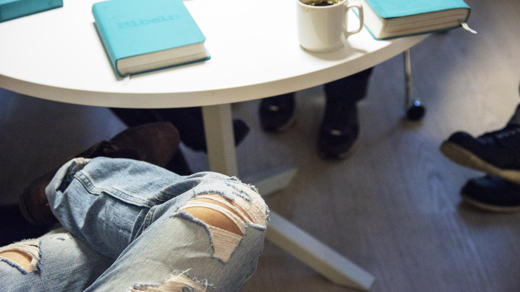Någon med trasiga jeans sitter med benen i kors vid ett bord med några andra. På bordet ligger några biblar.
