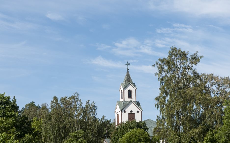 Ett kyrktorn sticker upp bakom trädkronorna mot blå himmel.