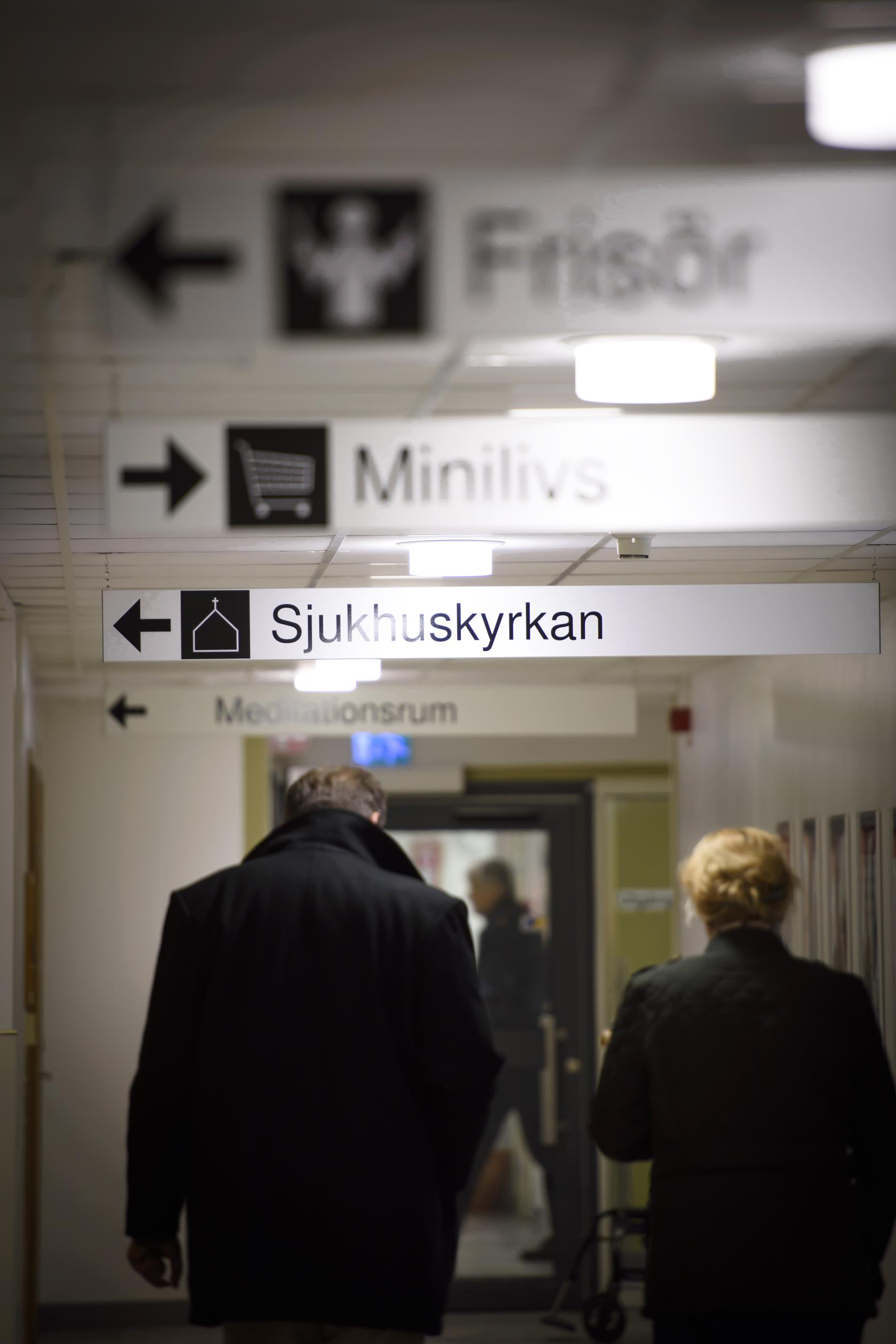 En man och en kvinna går i en korridor. I taket hänger olika skyltar med texterna Frisör, Minilivs och Sjukhuskyrkan.