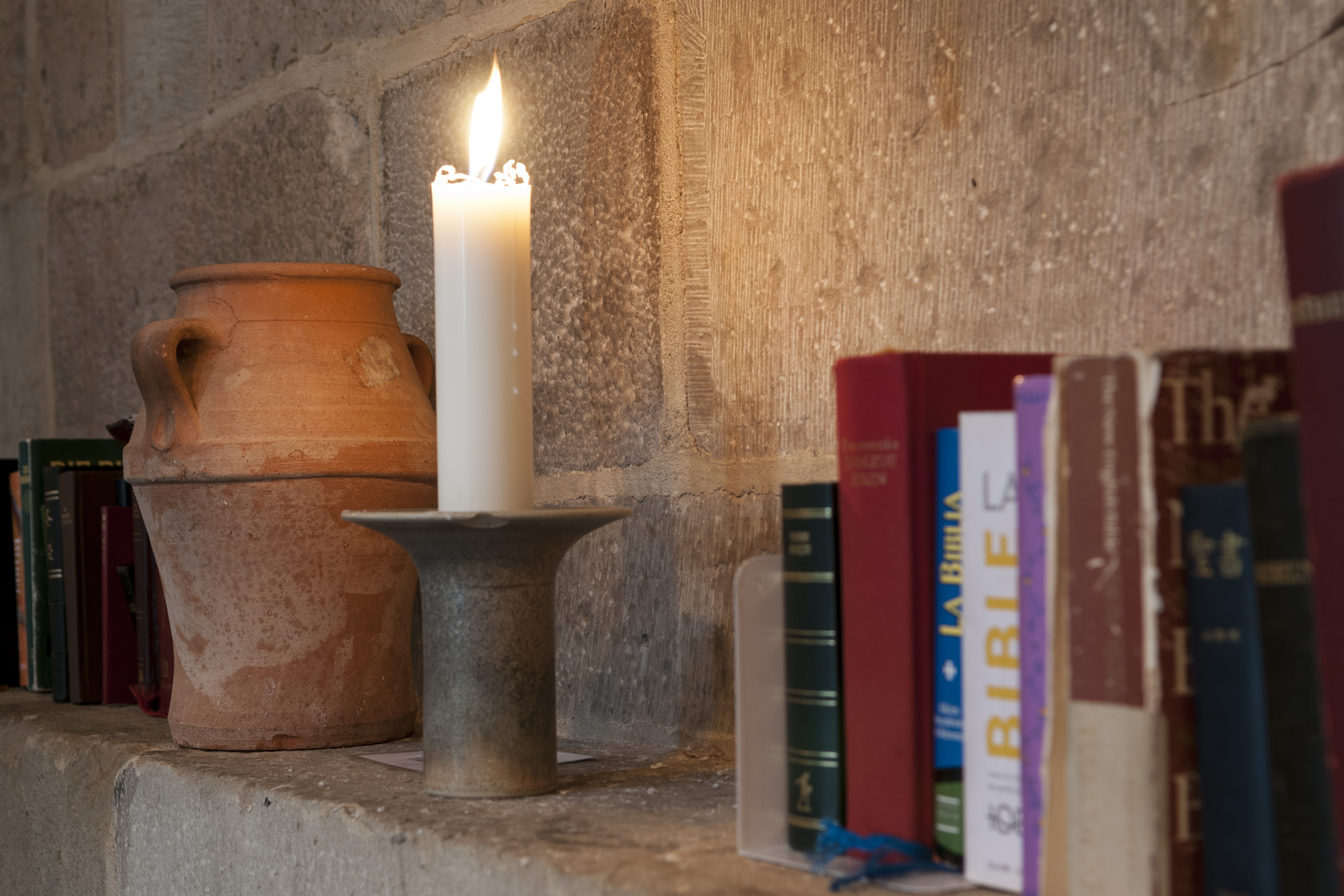 Biblar på olika språk, ett brinnande stearinljus och en kruka står på en stenhylla.