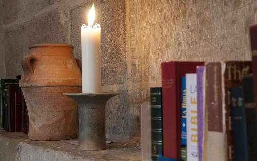 Biblar på olika språk, ett brinnande stearinljus och en kruka står på en stenhylla.