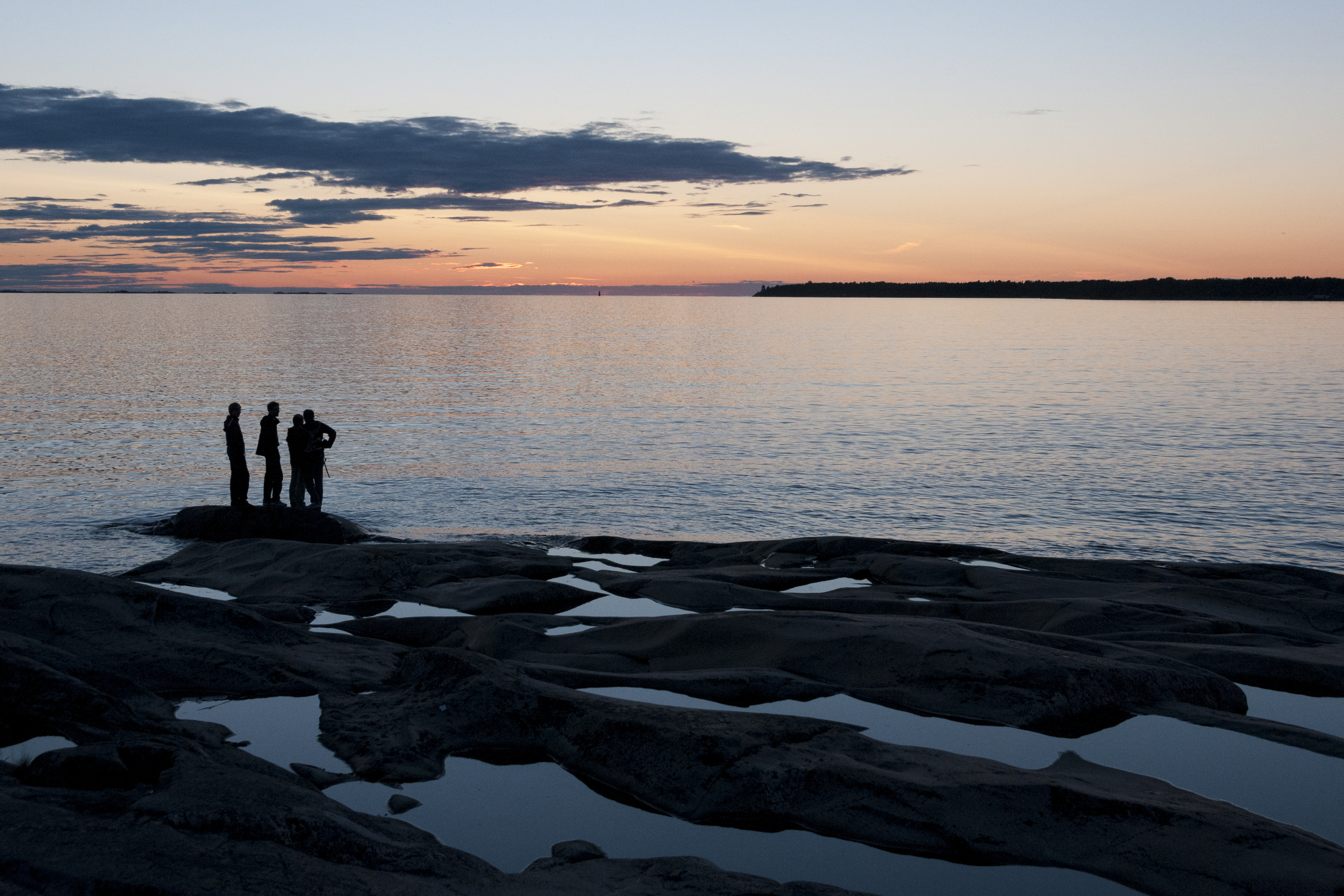Några människor står ute på klipporna vid vattnet i solnedgången.