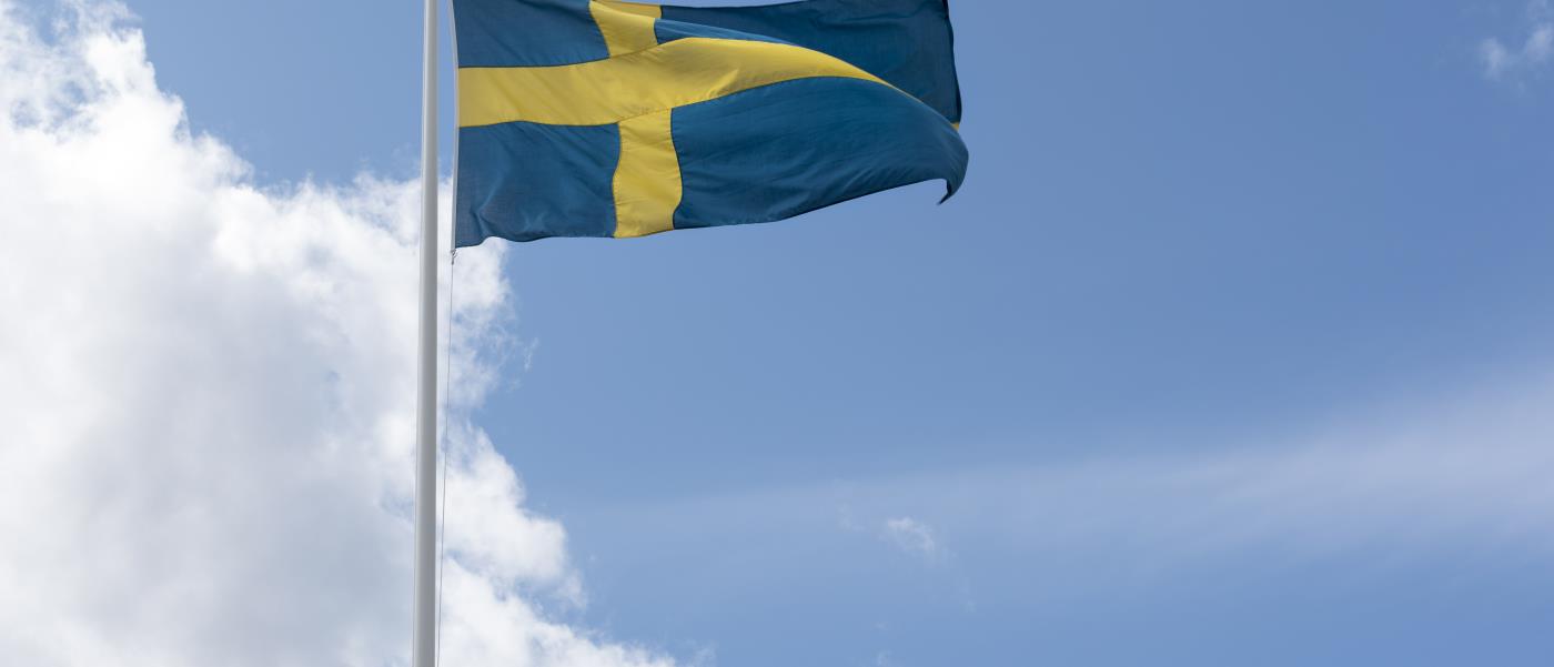 En flaggstång med svenska flaggan mot blå och molnig himmel.