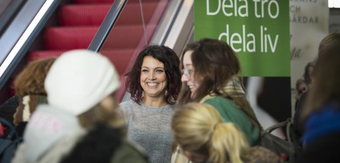 En grupp människor står bredvid en rulltrappa vid en grön skylt med texten Dela tro dela liv.