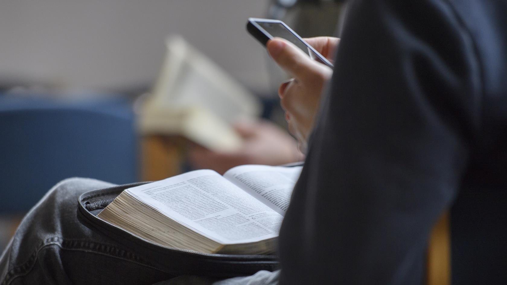 Någon sitter med en bok i knät och mobiltelefonen i handen.