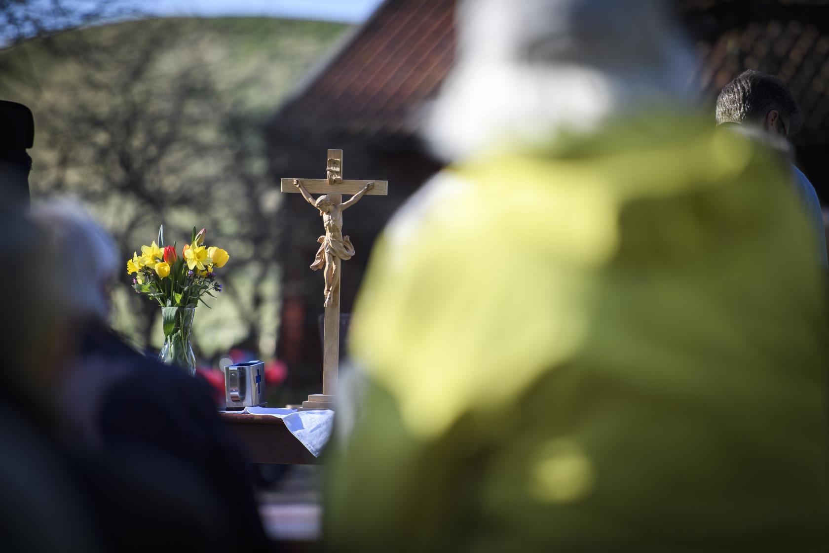 Ett krucifix i trä och en vas med påskblommor står på ett altare utomhus. Suddiga personer i förgrunden.