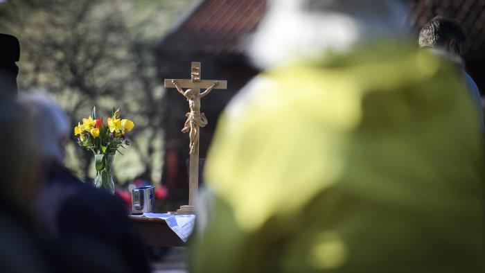 Ett krucifix i trä och en vas med påskblommor står på ett altare utomhus. Suddiga personer i förgrunden.
