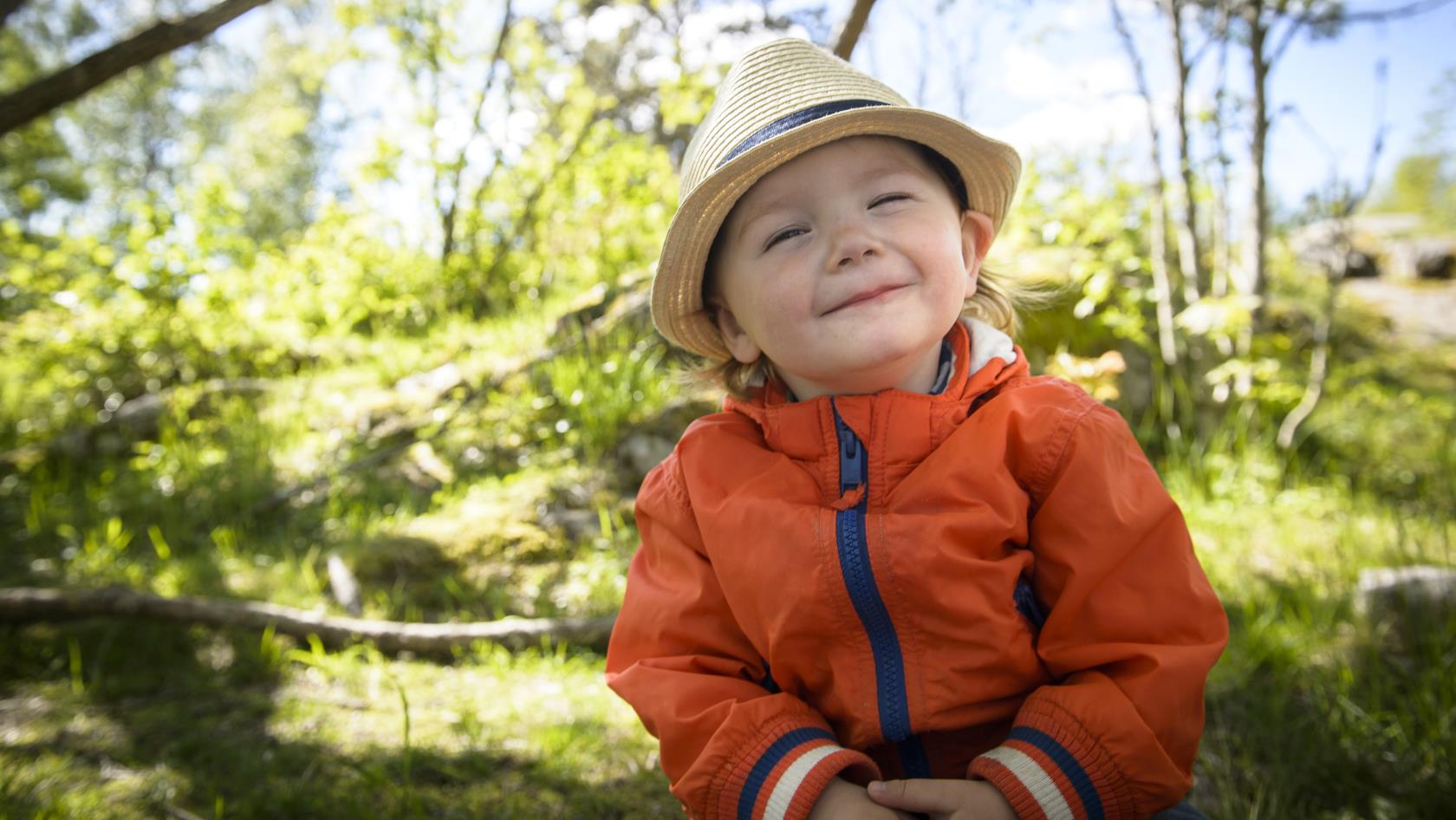 En liten pojke med halmhatt sitter ute i naturen och ler mot kameran.