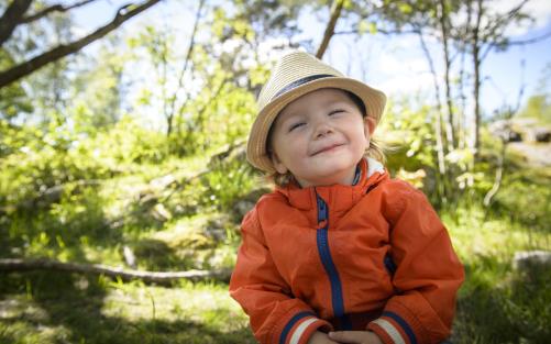 En liten pojke med halmhatt sitter ute i naturen och ler mot kameran.
