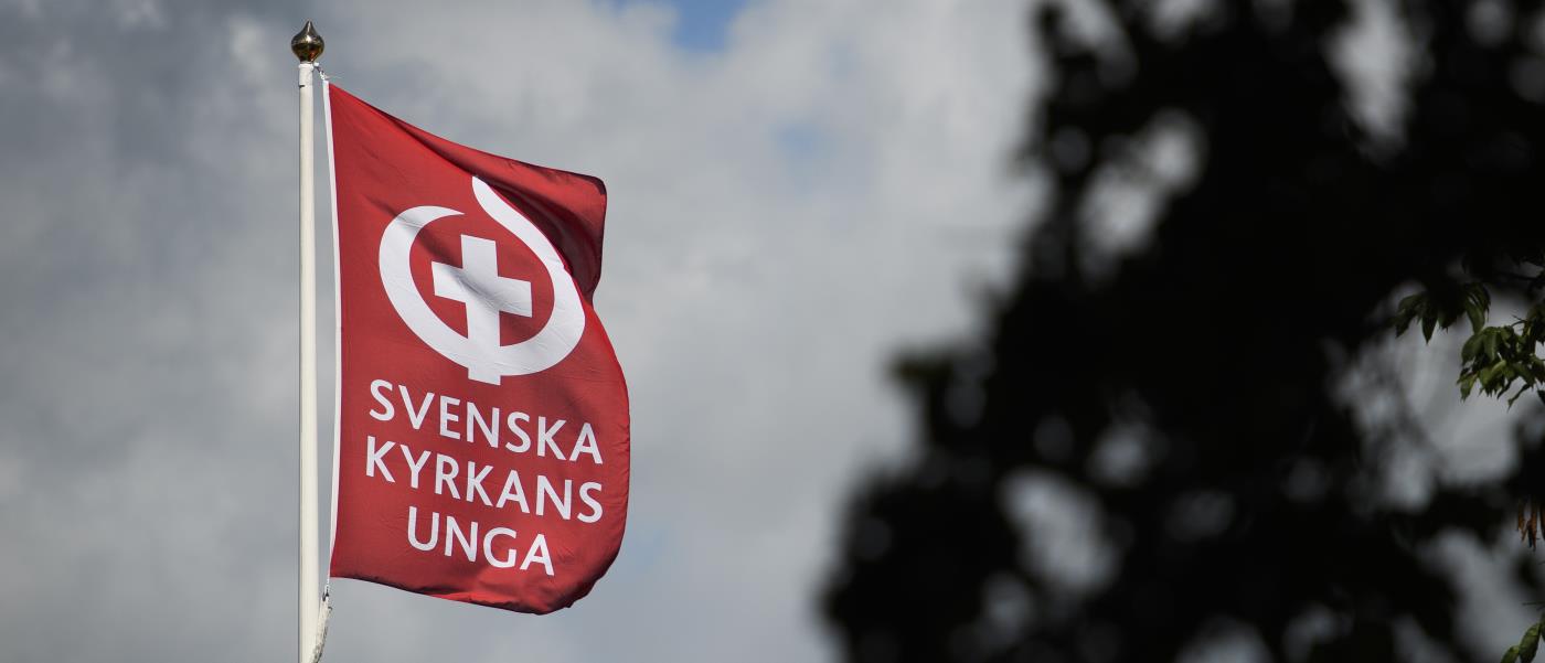 En flaggstång med en röd flagga med texten Svenska kyrkans unga och deras symbol.