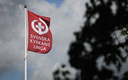 En flaggstång med en röd flagga med texten Svenska kyrkans unga och deras symbol.