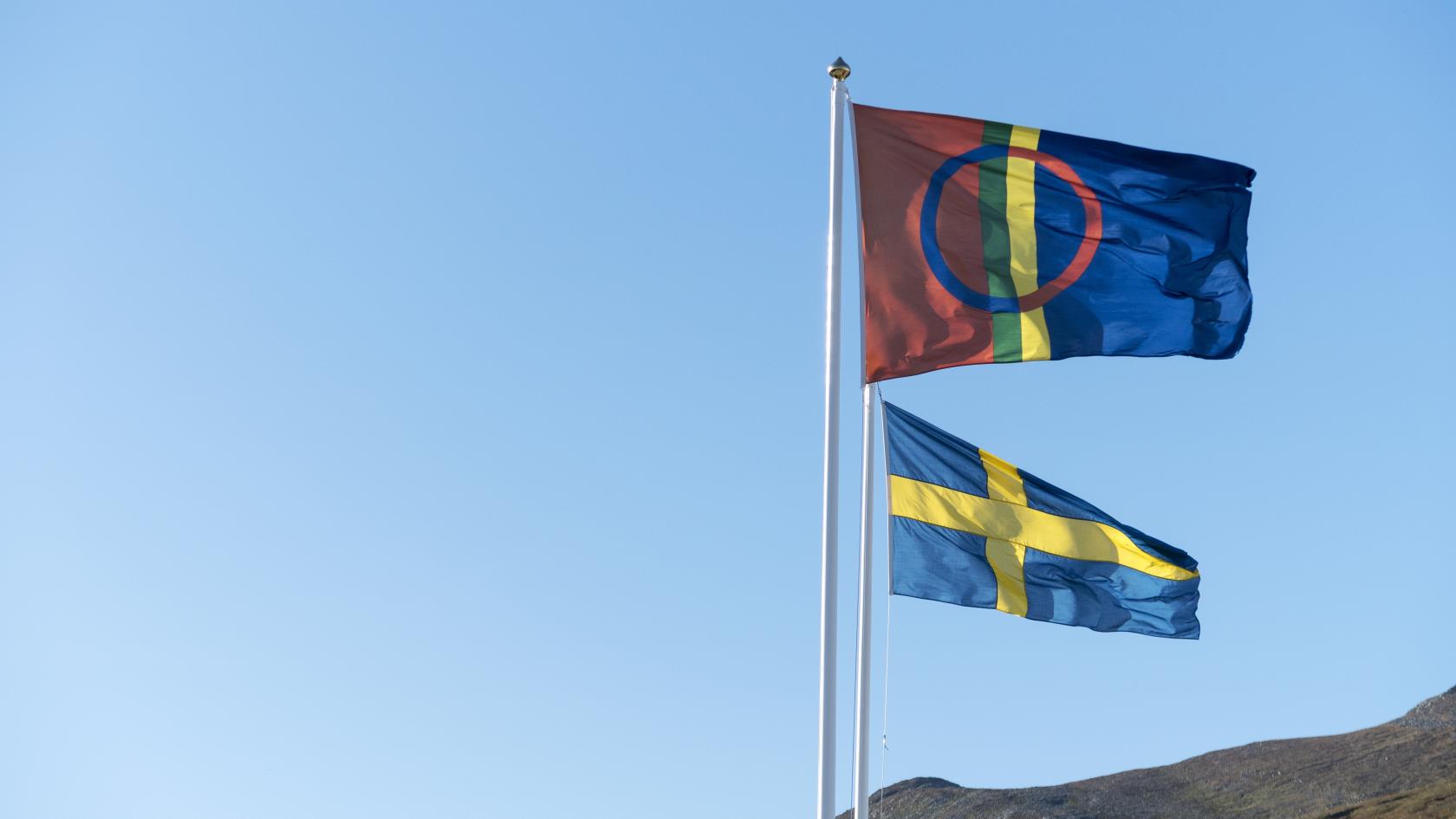 Två flaggstänger med samiska och svenska flaggan med blå himmel och berg i bakgrunden.