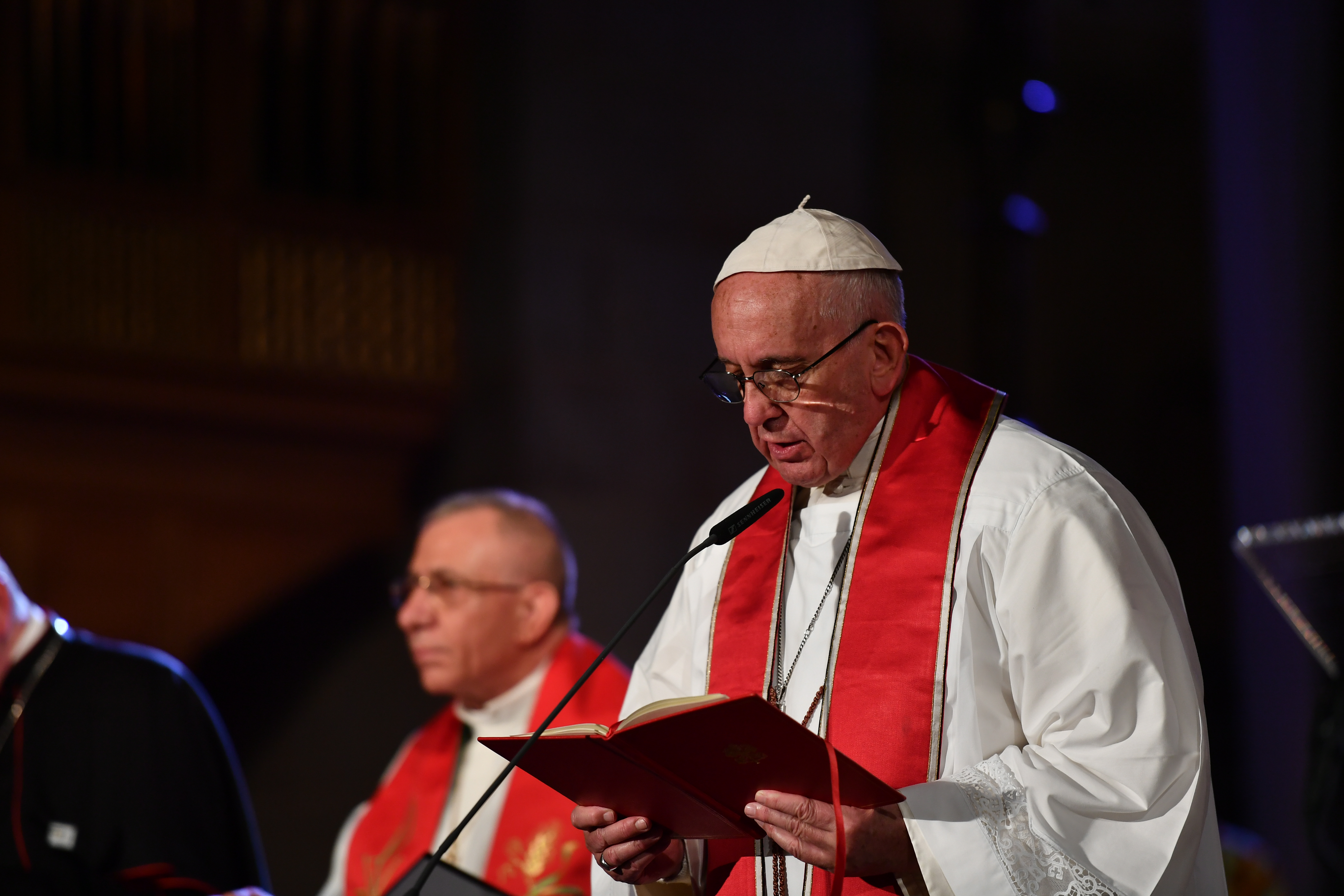Påve Franciskus i vit dräkt och röd stola läser högt ur en bok i en mikrofon.