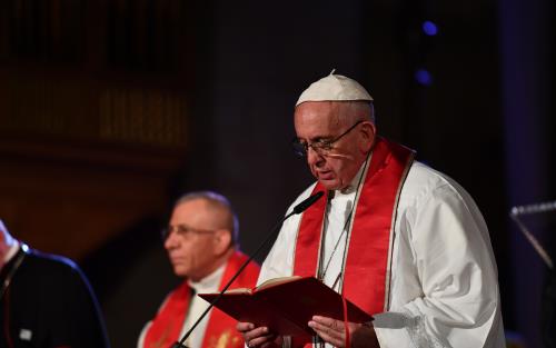 Påve Franciskus i vit dräkt och röd stola läser högt ur en bok i en mikrofon.