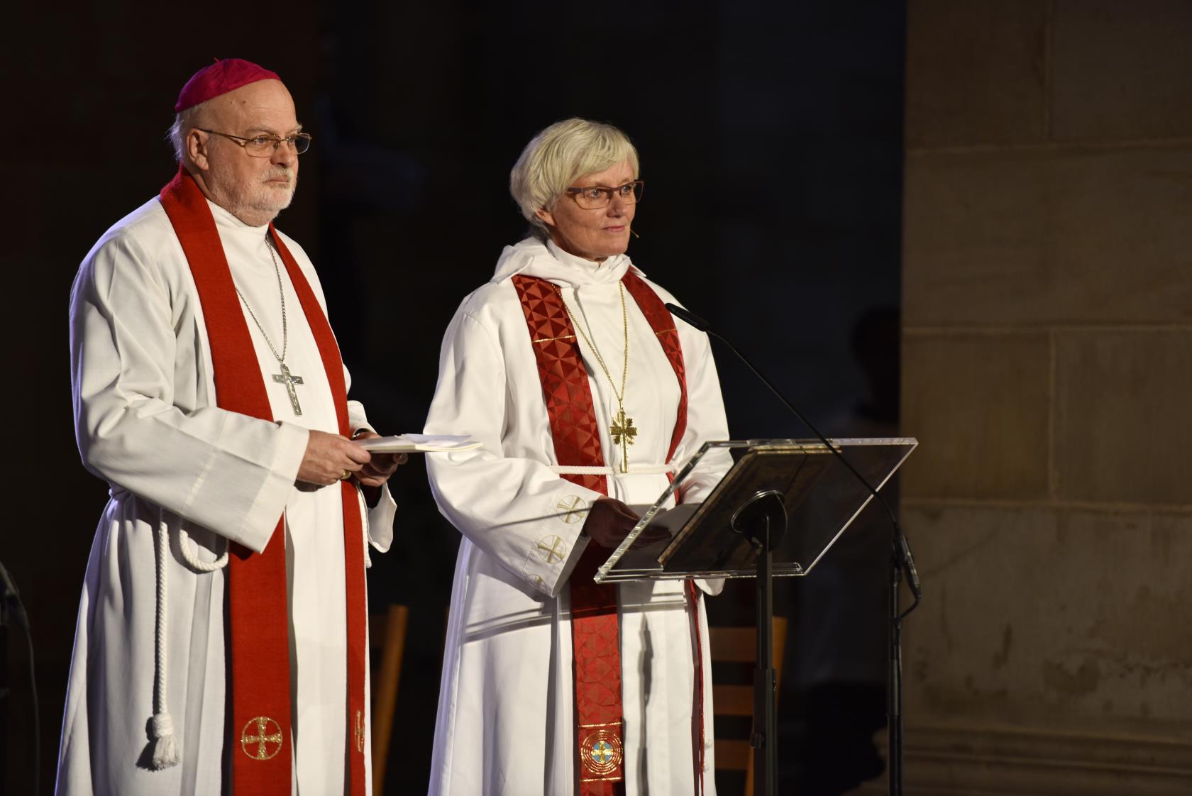 Biskoparna Anders Arborelius och Antje Jackelén står i strålkastarljus i en kyrka och talar i en mikrofon.