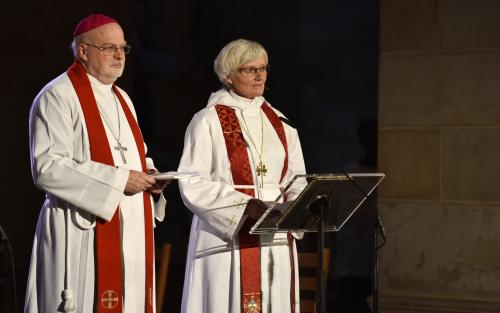 Biskoparna Anders Arborelius och Antje Jackelén står i strålkastarljus i en kyrka och talar i en mikrofon.