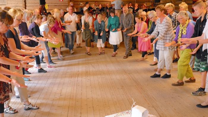 En grupp människor i ring håller händerna utsträckta framför sig. I mitten står ett litet bord med ett fat med oblater och en övertäckt kalk.