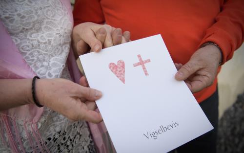 En man och en kvinna håller i ett papper med ett hjärta, ett kors och texten Vigselbevis.
