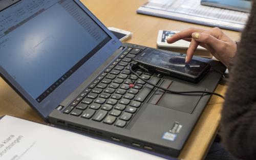 På en uppslagen laptop ligger en mobiltelefon som någon har sitt pekfinger på.