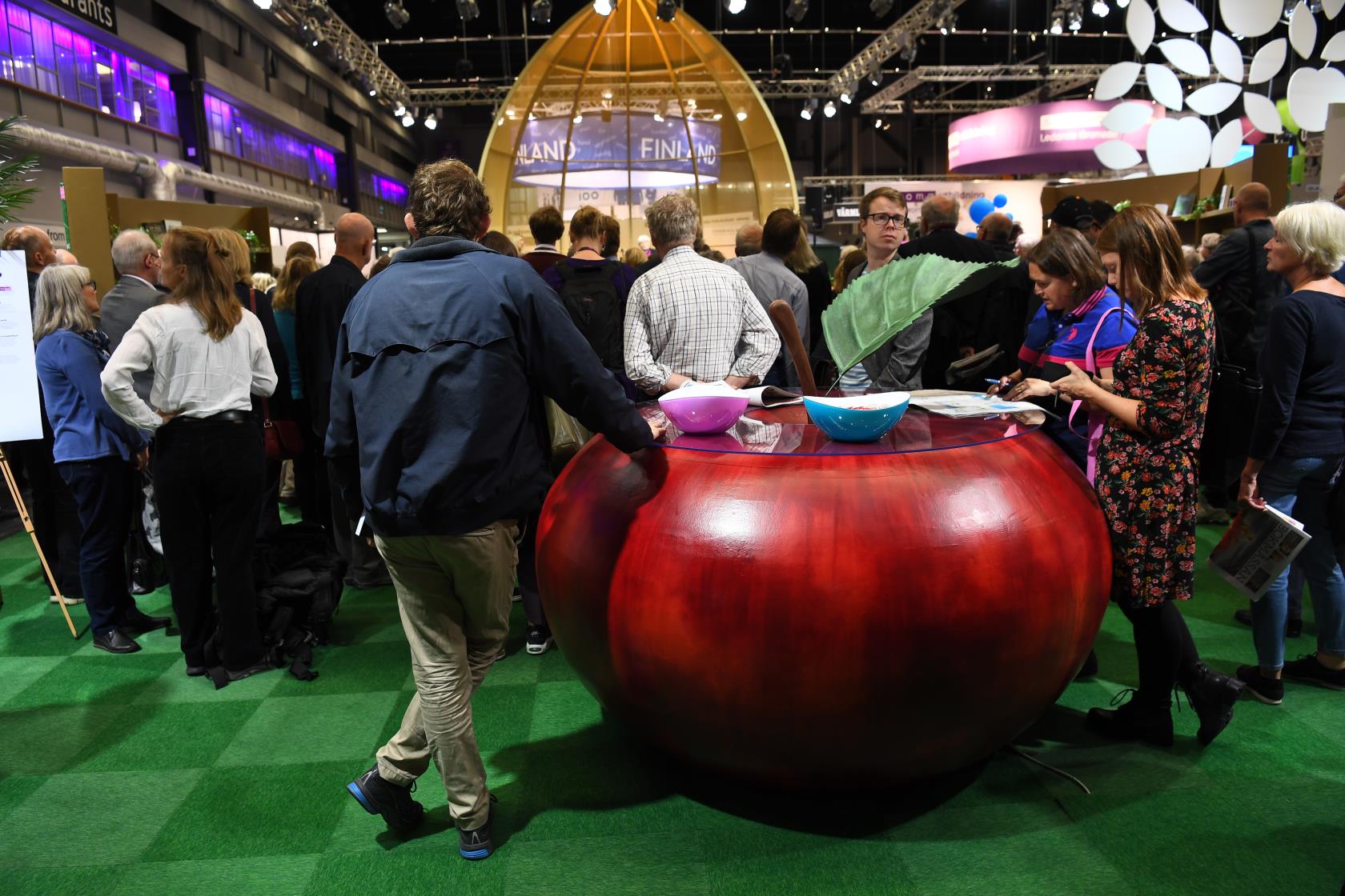 Mässdeltagare framför en fågelbur-liknande scen. I förgrunden ett runt bord i form av ett rött äpple.