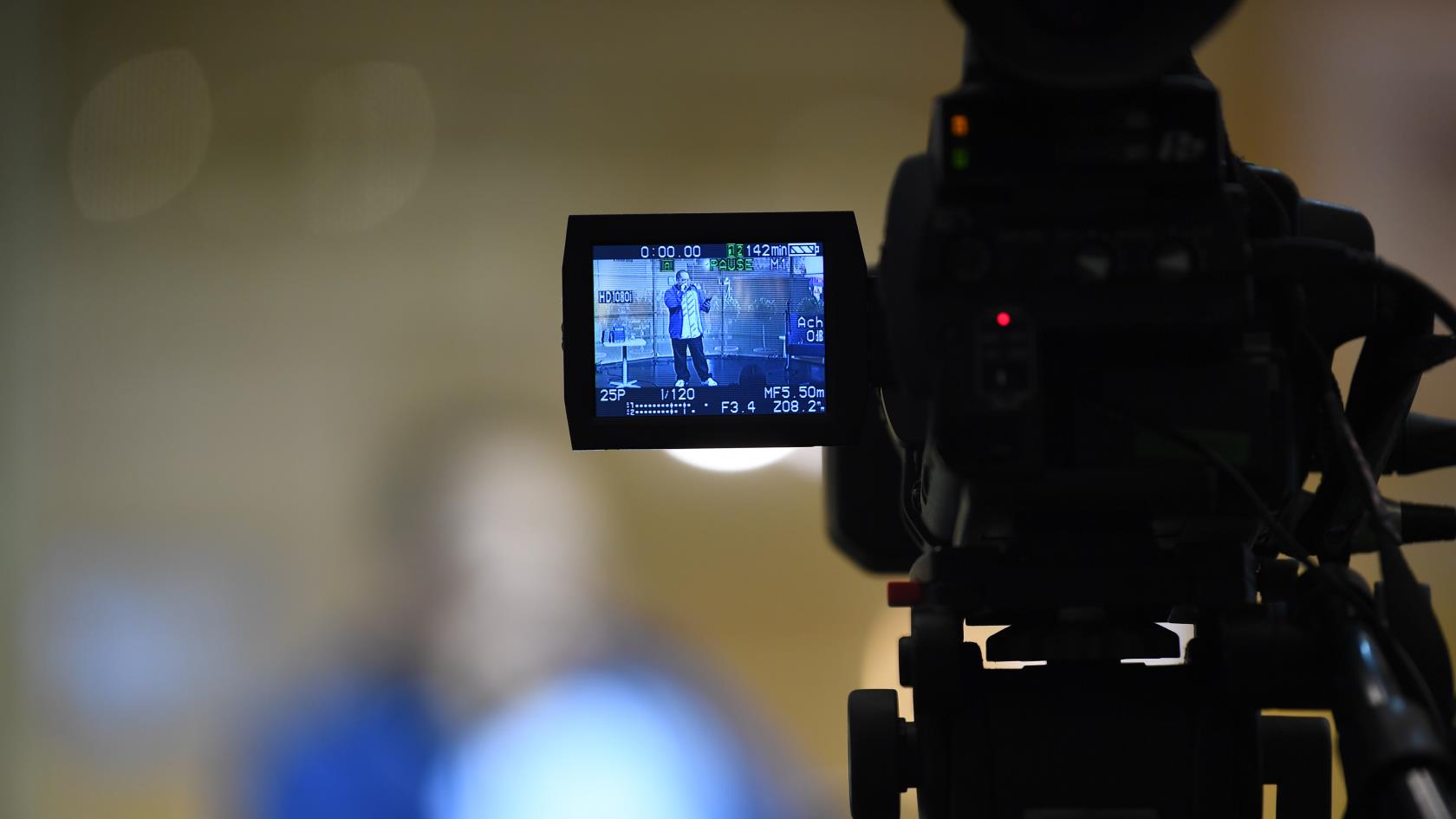 En filmkamera som spelar in. I kamerans display syns en man som står på en scen och talar.