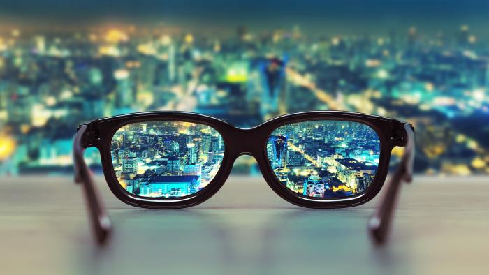 Ett par glasögon ligger i ett fönster med utsikt mot en storstad som lyser i kvällen. Staden är suddig utom genom glasögonen.