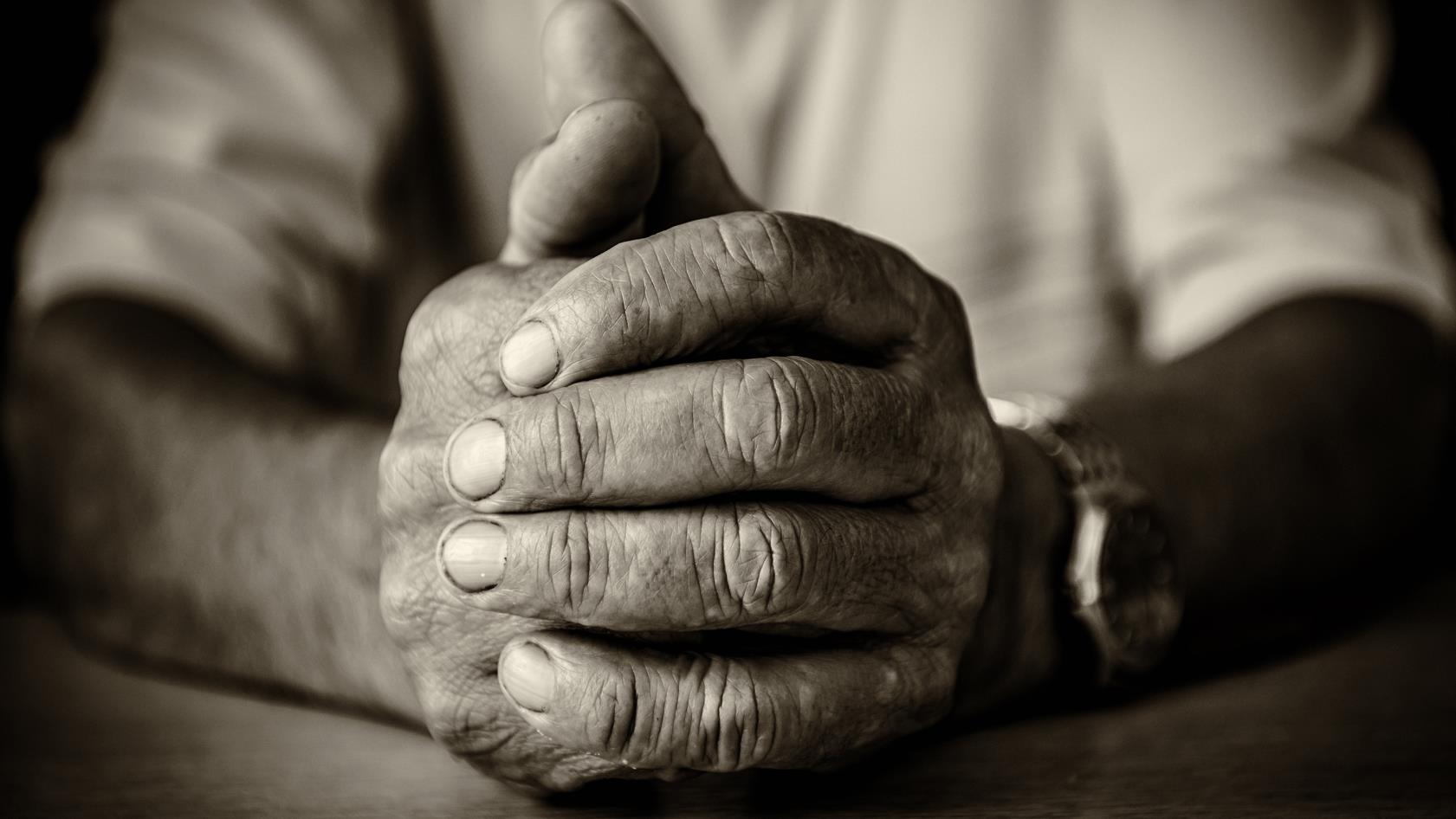 En svartvit bild på en gammal mans händer. Den högra handen håller om den vänstra. Händerna vilar mot ett bord.