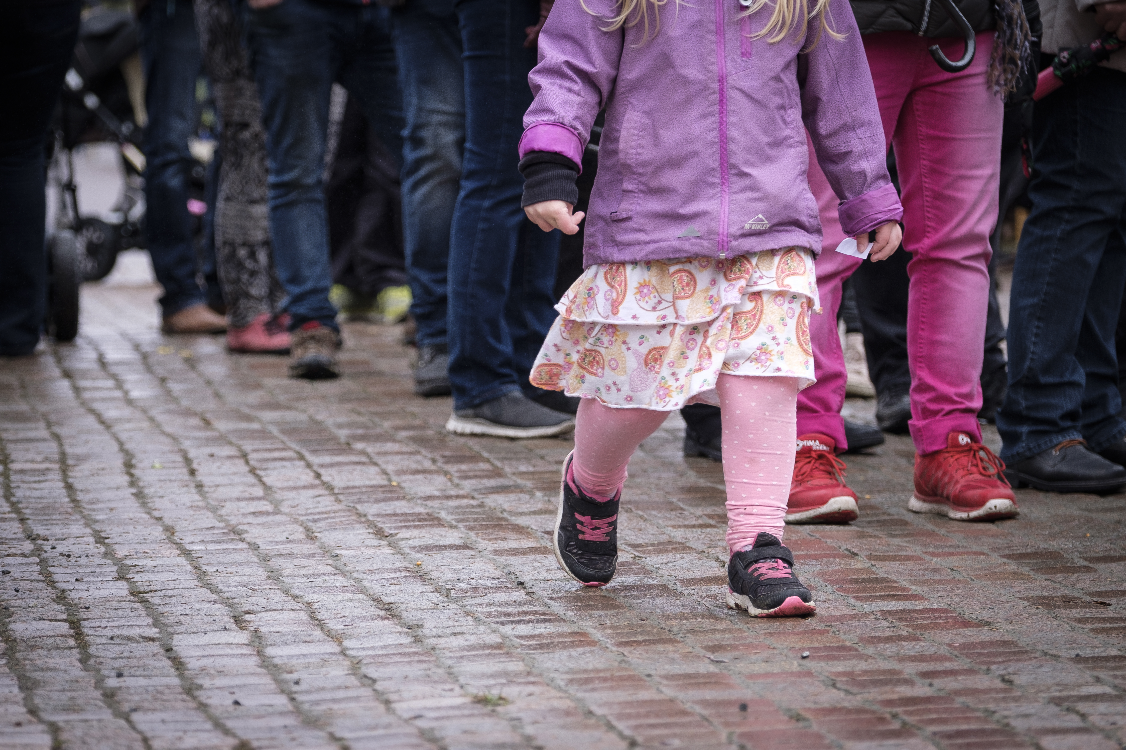 En liten flicka med rosa kläder går på en blöt kullerstensgata. En stor grupp människor syns bakom henne.